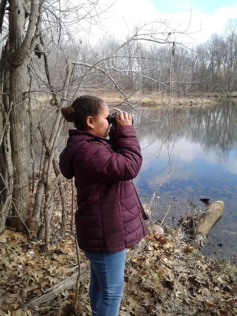 A young student is using binoculars to look at a nearby lake/woodland.