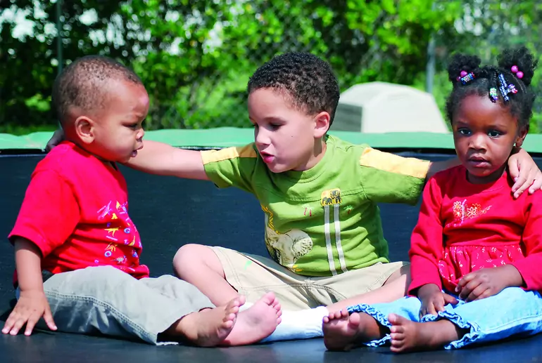 Three young children of varying ethnicities sitting on the ground looking unhappy. One child has his arms around the other two.