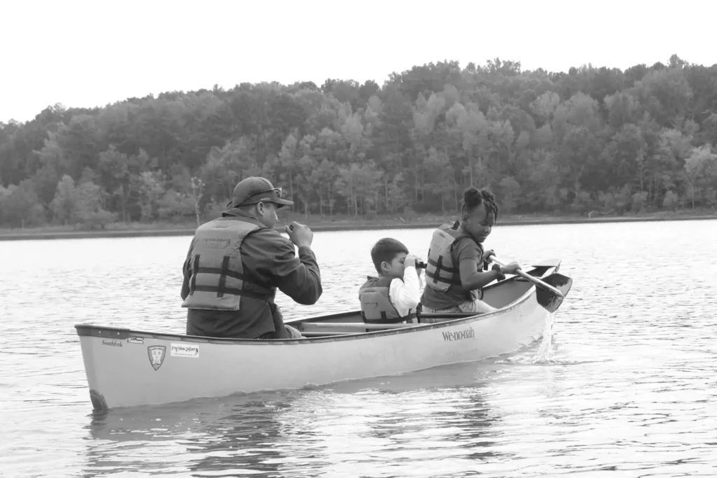 Two students and an adult in a canoe out on a lake. One student is paddling, the other is using binoculars to look at the water.