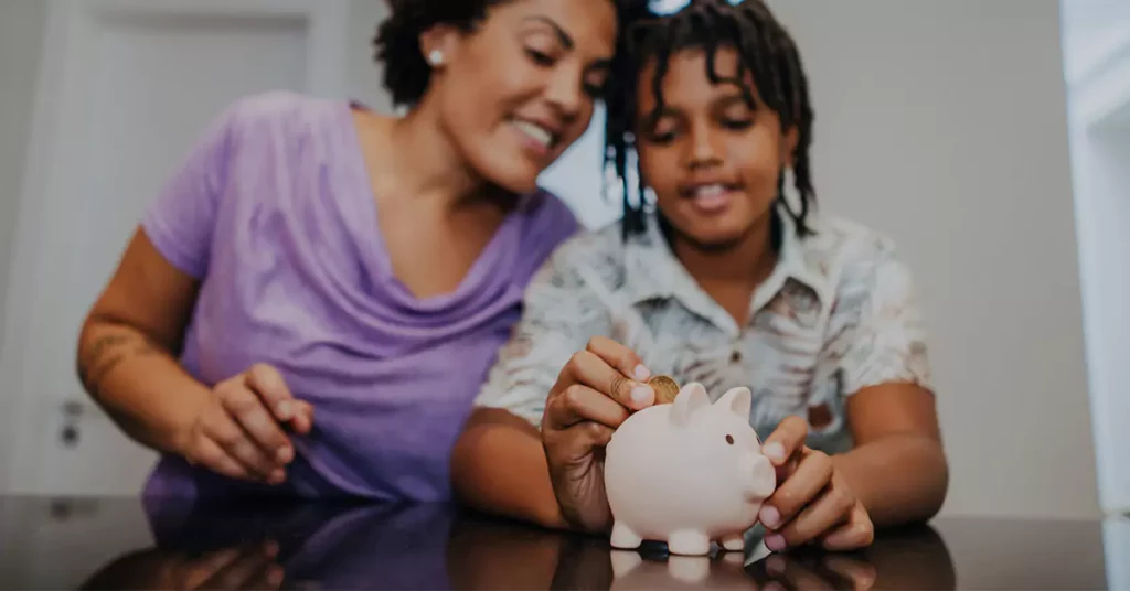 A mother watching her child put coins into a small piggy bank.