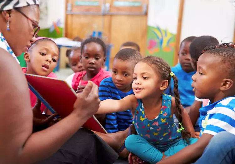 A group of young Black children watching as an adult reads to them.